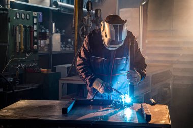 Kaynakçı fabrikadaki işyerinde kaynak yaparken kıvılcımlar uçuşurken koruyucu bir kask takıyor..