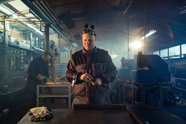 Tamirci, otomatik kaynak aletini işe hazırlarken meslektaşı arkada öğütme işiyle uğraşırken fark edilebilir..