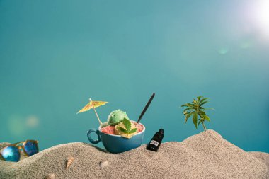 Bardakta dondurma, bir damla CBD yağı ve kumda bir dal marihuana. Yanında güneş gözlükleri, dondurma süslemeleri, deniz kabukları var..