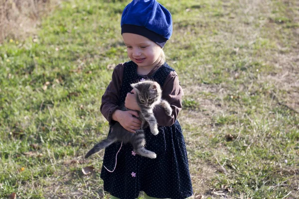 Kleines Mädchen in Kleid und Baskenmütze mit einem grauen Kätzchen in der Hand — Stockfoto