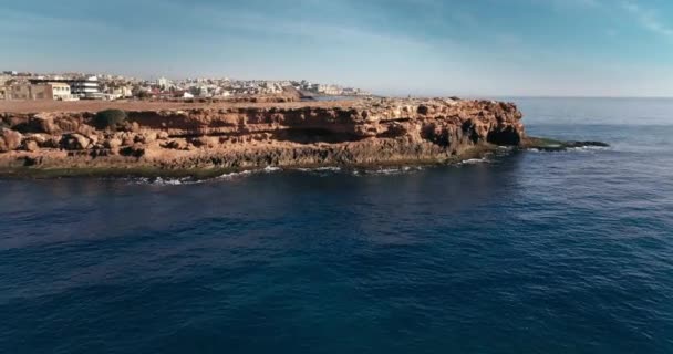 Drone si avvicina alle rocce e si affaccia sulla tranquilla costa rocciosa blu acqua di mare e il paesaggio urbano in lontananza. — Video Stock