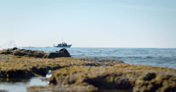 Рыбацкая лодка на Средиземном море воды плавает на горизонте в ясный день с волнами столкновения с камнями на переднем плане. — стоковое видео