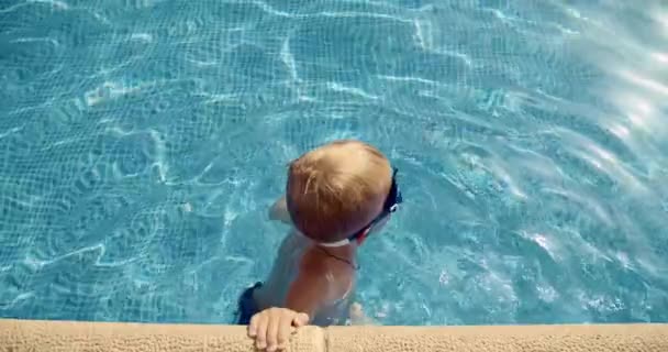 Ung pojke lär sig simma i poolen en solig sommardag med glasögon i klarblått vatten — Stockvideo