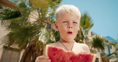 Küçük mutlu çocuk güneşli bir günde dışarıda büyük bir dilim karpuz yiyor ve gülümsüyor.