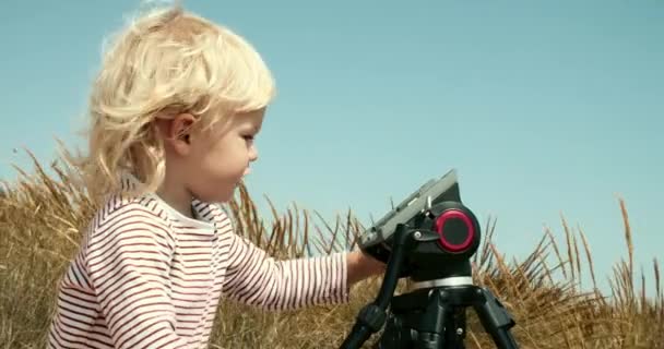 Little caucasian boy with blonde hair plays with fathers tripod outdoors — Stock Video