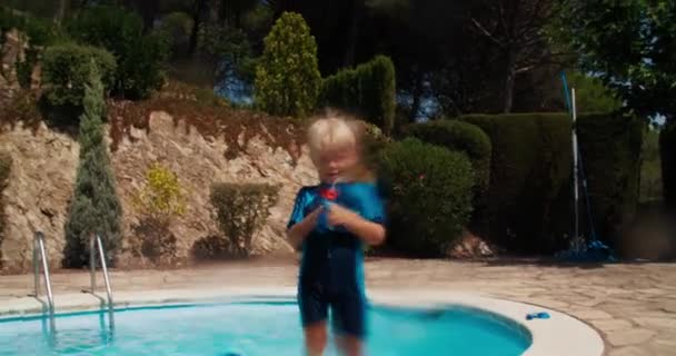 Cute baby boy shooting with water gun directly to camera standing next to pool — Stock Video