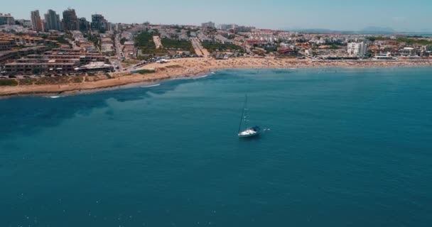 Дрон летает вокруг парусной яхты с видом на пляж и город в летний день — стоковое видео