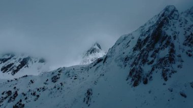 Soğuk kış manzarası, alçak bulutlarla çevrili karlı zirveye odaklanmış dağların üzerinden uçuyor. Donmuş kırsal manzara. 