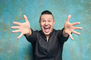 Kırmızı pantolon ve siyah gömlek, tıraşlı tapınaklar ve kaygan arka saç ile saç modeli, mavi duvar, sakal, parlak gülümsüyor ve kollarını öne çeker genç bir adamın portresi