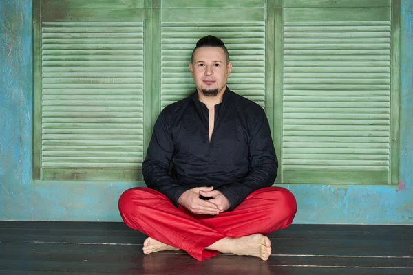 Portrait of a young man, black shirt and red slacks, hairstyle with shaved temples and slicked- back hair at the top of the head, different emotions, green wall, brown floor, beard, lotus posture