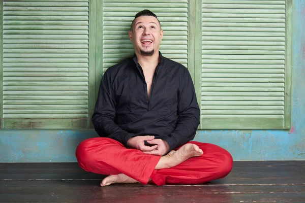 Retrato de un joven, camisa negra y pantalones rojos, peinado con sienes rasuradas y cabellos en la parte superior de la cabeza, diferentes emociones, pared verde, piso marrón, barba, postura de loto — Foto de Stock