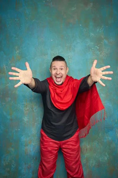 Portrait of a young man in red slacks and a black shirt, hairstyle with shaved temples and slicked- back hair at the top of the head, blue wall, beard, bright smiles and pulls the arms forward — Stock Photo, Image