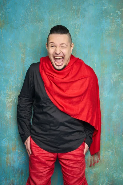 Portrait of a young man in red slacks and a black shirt, hairstyle with shaved temples and slicked- back hair at the top of the head, blue wall, beard, bright smiles and pulls the arms forward — Stock Photo, Image