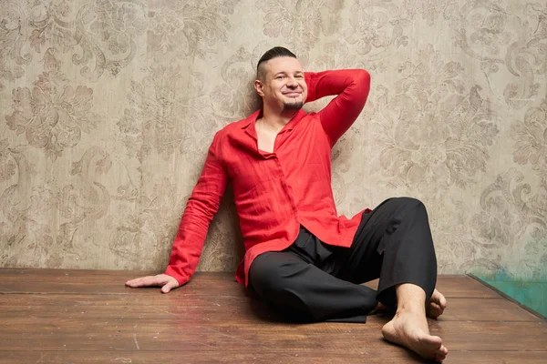 Portrait of a young man, in black slacks and a red shirt, hairstyle with shaved temples and slicked- back hair at the top of the head, emotions, beige wall with drawings, wood brown floor, beard