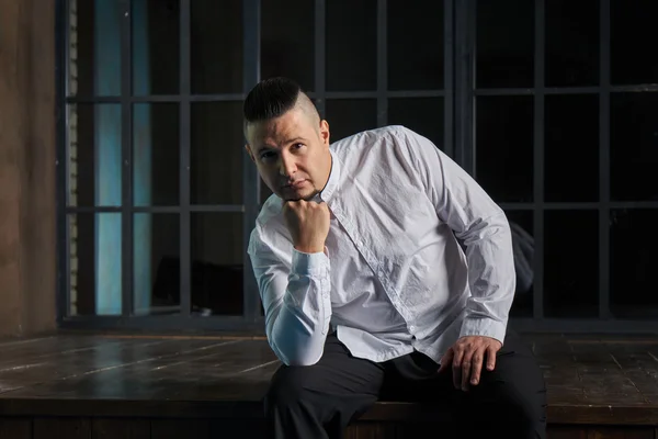 Portrait of a young man, in black slacks and a white shirt, hairstyle with shaved temples and slicked- back hair at the top of the head, different emotions, black window, brown floor, Brick wall, beard, skinhead, brutal, evil, killer