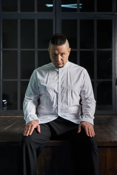 Portrait of a young man, in black slacks and a white shirt, hairstyle with shaved temples and slicked- back hair at the top of the head, different emotions, black window, brown floor, Brick wall, beard, skinhead, brutal, evil, killer