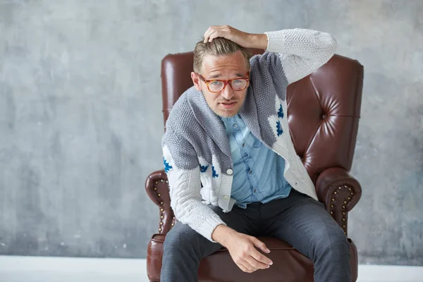 Retrato de un hombre inteligente con estilo con gafas mira a la cámara, buena vista, pequeño sin afeitar, carismático, camisa azul, suéter gris, sentado en una silla de cuero marrón, agarra su cabeza — Foto de Stock