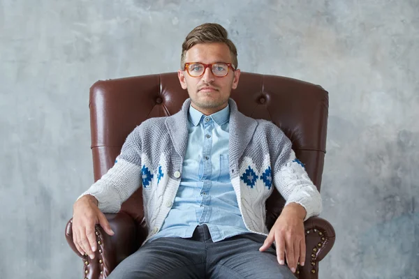 Retrato de un hombre inteligente con estilo con gafas mira a la cámara, buena vista, pequeño sin afeitar, carismático, camisa azul, suéter gris, sentado en una silla de cuero marrón —  Fotos de Stock