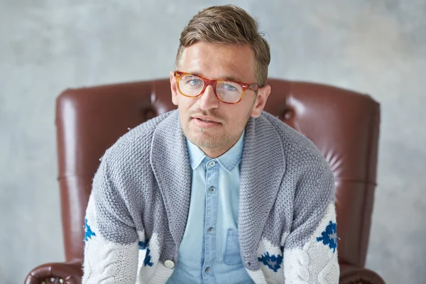 Retrato de un hombre inteligente con estilo con gafas mira a la cámara, buena vista, pequeño sin afeitar, carismático, camisa azul, suéter gris, sentado en una silla de cuero marrón —  Fotos de Stock