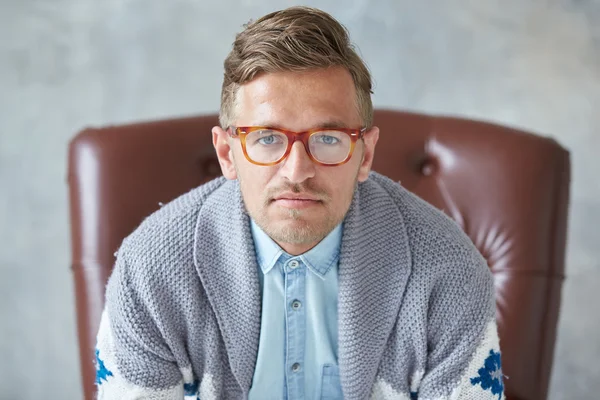 Retrato de un hombre inteligente con estilo con gafas mira a la cámara, buena vista, pequeño sin afeitar, carismático, camisa azul, suéter gris, sentado en una silla de cuero marrón —  Fotos de Stock