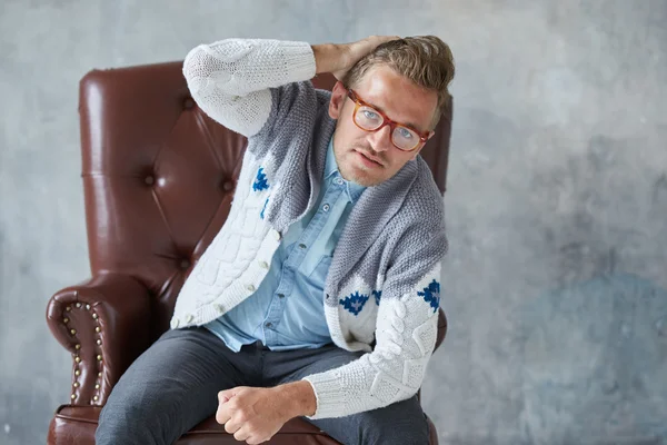 Retrato de un hombre inteligente con estilo con gafas mira a la cámara, buena vista, pequeño sin afeitar, carismático, camisa azul, suéter gris, sentado en una silla de cuero marrón, agarra su cabeza —  Fotos de Stock