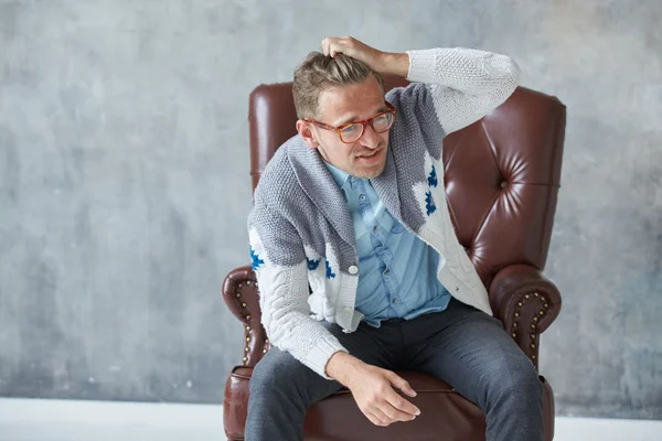 Retrato de un hombre inteligente con estilo con gafas mira a la cámara, buena vista, pequeño sin afeitar, carismático, camisa azul, suéter gris, sentado en una silla de cuero marrón, agarra su cabeza —  Fotos de Stock