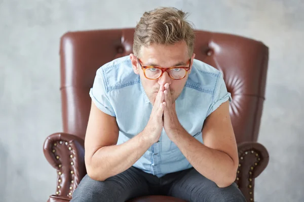 Retrato de un hombre elegante con gafas, mira a la cámara, sin afeitar, carismático, camisa azul con manga corta, sentado en una silla de cuero marrón, diálogo, negociación, manos como en la oración — Foto de Stock