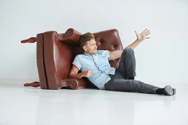 Brutal hombre acostado junto a un sillón de cuero marrón, sillón acostado en su lado en el suelo, un hombre toma de la mano detrás de la silla — Foto de Stock