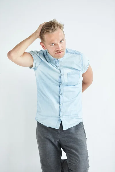 Hombre brutal con una barba pequeña se apoya en la pared, con una camisa azul de manga corta y pantalones grises, las manos están detrás de su espalda — Foto de Stock