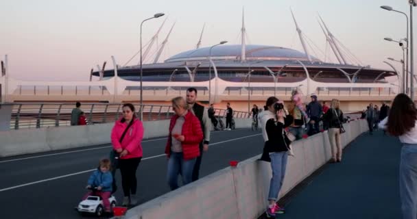 Ryssland, St Petersburg, 01 September 2020: Människor går på bron vid solnedgången under pandemin av covid-19, rida elektroskateboards, gör bilden, arenan Gazprom är på bakgrunden — Stockvideo