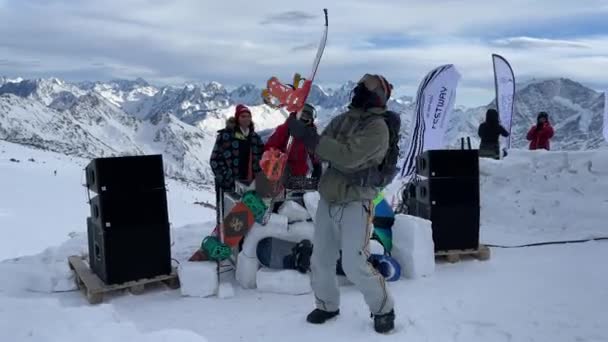 Oroszország, Kabardino-Balkariya, 2021. január 07.: A DJ az Elbrus-hegy legnépszerűbb pontján játszik, a lány a közelben táncol, a snowboardos táncol, a hegy a háttérben, Azaus síközpont — Stock videók