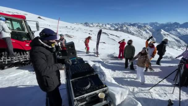 Oroszország, Kabardino-Balkariya, 2021. január 07.: A DJ az Elbrus-hegy legnépszerűbb pontján játszik, a lány a közelben táncol, mögöttük a snowboardos, a háttérben a hegy, Azaus síközpont — Stock videók