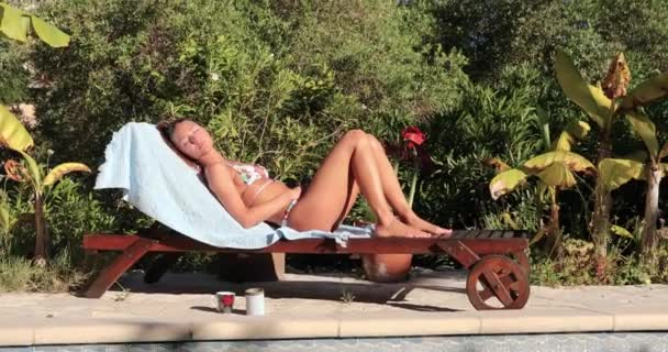 A fiatal lány egy gyönyörű test, harmonikus, fekszik egy fa kanapé társalgó és napfürdők a napon, közel a medence, a külön fürdőruha, egy csésze kávét, a háttérben zöld növényzet — Stock videók