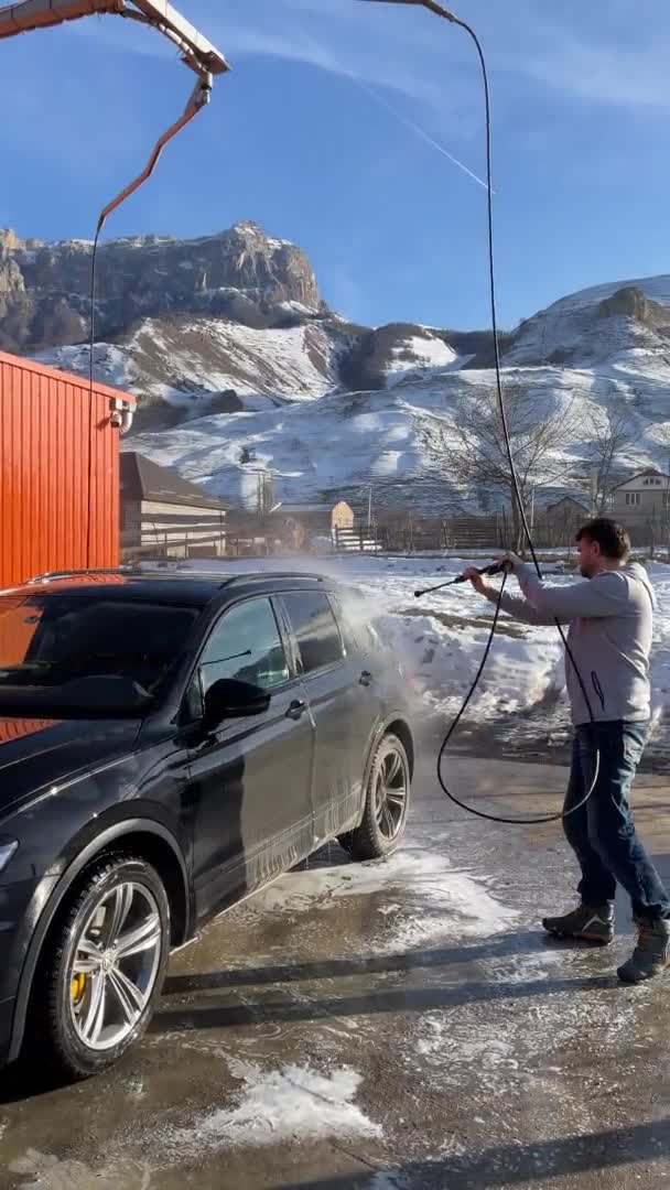 Россия, Кабардино-Балкария, 05 января 2021 года: Молодой человек моет машину на автомойке в солнечное морозное утро, брызги разбросанные по разным сторонам, заснеженные горы на заднем плане — стоковое видео