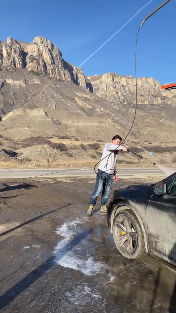 Russie, Kabardino-Balkariya, 05 janvier 2021 : Le jeune homme lave la voiture sur le lavage de voiture dans le matin givré solaire, éclaboussures disperser dans les différentes parties, montagnes enneigées sur un fond — Video