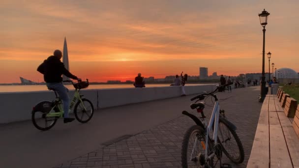 Russland, St.Petersburg, 10. Juni 2020: Menschen beobachten einen unwahrscheinlichen Sonnenuntergang über dem höchsten Wolkenkratzer Europas, dem Gazprom Lakhta Center, dem überfüllten Damm, Menschen fahren Fahrrad — Stockvideo