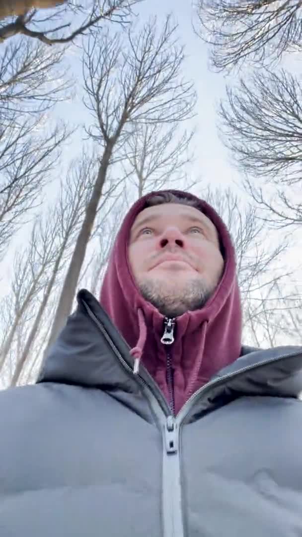 Egy fiatal férfi sétál a parkban, kapucnis pulóvert és kabátot visel, a fiatalember maga készít egy videót, néha a kamerába néz, levelek nélküli fák a háttérben. — Stock videók