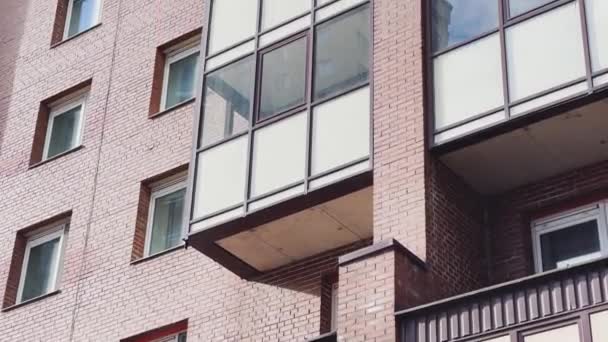 Facade of the high-floor house with the glazed balconies, the bottom view up, slow movement of a video camera, brick color, cloudy weather, a huge housing estate — стоковое видео