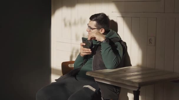 Russie, Saint-Pétersbourg, 11 avril 2021 : L'adolescent sérieux est assis fonctionne dans le smartphone, il est assis au café sur le soleil et il attend le café, téléphone dans une couverture de couleur verte — Video