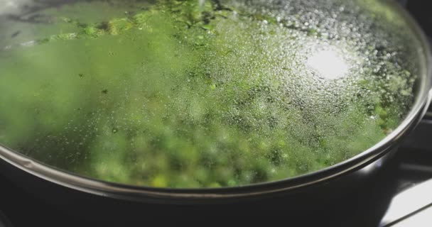 Свіжий зелений горох обсмажується на сковороді через скляну кришку, фокус на краплі води на скляній кришці, яскрава і дієтична страва, здоровий спосіб життя — стокове відео