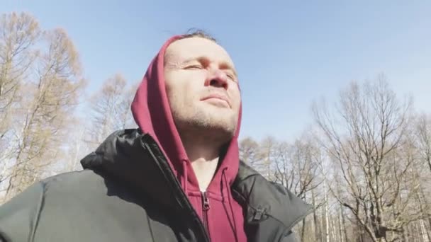 The handsome men is walking in the park at sunny day, is wearing a sweatshirt with a hood and a down jacket, sometimes look at the camera, trees without leaves in the background — Stock Video