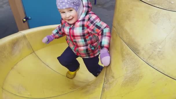 Russland, St.Petersburg, 11. April 2021: Das kleine Mädchen spielt auf einem Spielplatz, das Mädchen trägt warme Kleidung, trübes Wetter, sie ist glücklich und fröhlich — Stockvideo