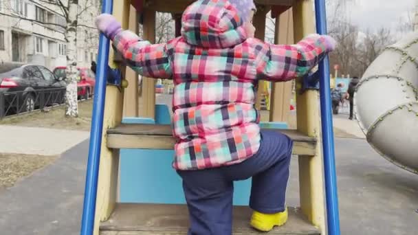 A menina brinca em um playground, a menina está vestida com roupas quentes, tempo nublado, ela feliz e alegre — Vídeo de Stock