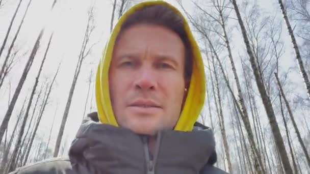 The handsome men is walking in the park at sunny day, is wearing a sweatshirt with a hood of yellow color and a down jacket, look at the camera, trees without leaves in the background — Stock Video