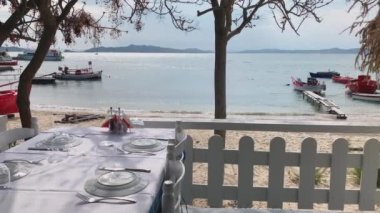 Boş bir halk plajı olan mavi bir göldeki restoran masasının manzarası gün doğumunda ağaçların arasından, turistler, masmavi sular ve balıkçı tekneleri olmadan arka planda dağlar.