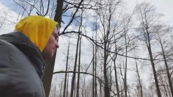 Les beaux hommes marchent dans le parc à la journée ensoleillée, portent un sweat-shirt avec une capuche de couleur jaune et une veste en duvet, caméra sur le côté droit, des arbres sans feuilles en arrière-plan — Video