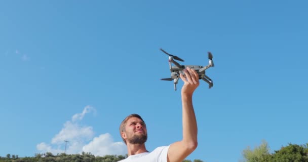 Pohledný chlapík spustí dron z rukou, Dolní pohled na létající dron proti modré obloze za slunečného počasí, Ruce sáhnou po dronu, senzorech a kameře, čepele v pohybu, záběry zblízka — Stock video