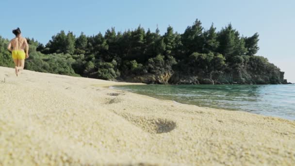 Slow motion beelden van de man in gele shorts en een naakte romp loopt op een verlaten strand, hij beweegt weg van een camera, het schiereiland met bomen op een achtergrond, het onderste uitzicht, geen mensen — Stockvideo