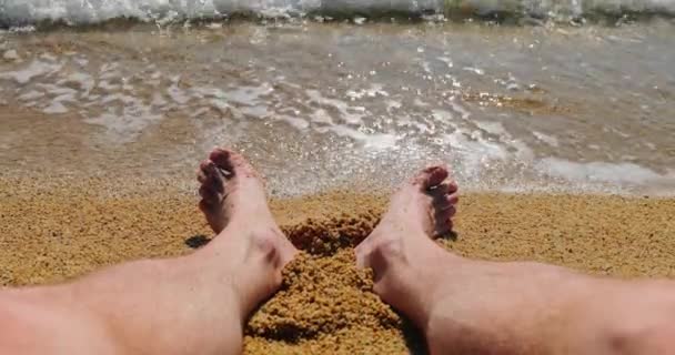 Las vacaciones mucho tiempo esperadas, el turista se sienta en la playa en la arena y moja los pies en el agua de mar, el agua azul lava los pies, las olas, el reflejo solar — Vídeos de Stock