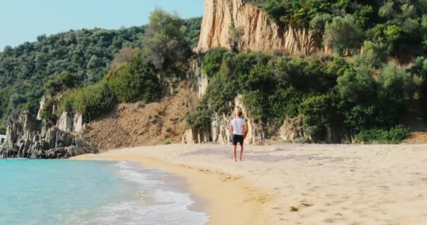 Slow motion beelden van de man in blauwe shorts en een wit t-shirt loopt op een verlaten strand, hij beweegt zich weg van een camera, de berg op een achtergrond, geen mensen — Stockvideo